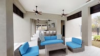 This single-story amenity facility is thoughtfully designed to offer residents a range of shared spaces that balance comfort and functionality. The covered outdoor lounge is a standout feature, offering an expansive, open-air gathering area with smooth stucco-finished walls, soaring ceilings, and multiple ceiling fans that ensure airflow and comfort. The space is furnished with wicker seating and vibrant blue cushioned sofas arranged around a low coffee table, creating an inviting setting for relaxation or socializing. The open-column layout allows the lounge to extend deeper into the structure, where a bar-height counter with seating and wall-mounted televisions provides additional entertainment options.

The laundry facility is a generously proportioned, high-ceilinged room with exposed steel beam framing and industrial-style lighting. A long row of commercial-grade washing machines lines one wall, with dryers mounted above, offering ample capacity. A wide folding counter runs along the opposite side, and polished concrete flooring ties the utilitarian aesthetic together. Windows and an exterior door provide natural light and ventilation.

A separate waiting or utility area features the same polished concrete floors and exposed overhead ductwork, with bench seating and neutral walls—a functional transitional space within the facility.