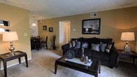 This single-floor space offers an open-concept layout that connects the living and dining areas in a seamless, functional arrangement. The living area is anchored by plush wall-to-wall carpeting in a neutral tone, providing a warm and comfortable foundation. Warm tan-hued walls create an inviting atmosphere, complemented by decorative framed artwork that adds visual interest. A textured ceiling adds subtle dimension to the room overhead, while a centrally located passageway leads to additional areas of the unit, contributing to the overall flow of the space.

The dining area is situated adjacent to the living room and is illuminated by a hanging pendant light fixture, adding a touch of elegance to the shared open space. The two areas are distinguished by their respective furnishings rather than physical dividers, reinforcing the open and connected feel of the layout. Ceiling-mounted HVAC vents throughout indicate a central air system. The overall ambiance is cohesive and well-organized, with a design that balances practicality and comfort in a compact, single-level setting.