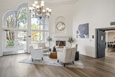 Located at 12200 Valley View St in Garden Grove, California, this expansive single-floor facility offers a generously sized, thoughtfully designed space suited for community gatherings and meetings. The grand living room serves as the centerpiece of the space, boasting dramatic double-height ceilings adorned with a wrought-iron chandelier and large arched windows that flood the area with natural light and provide views of the surrounding landscape. A classic white fireplace with an arched niche above serves as a refined focal point, complemented by light hardwood flooring, neutral tones, and elegant furnishings that create a welcoming and sophisticated ambiance. An adjacent seating area continues the warm, residential aesthetic with blue accent chairs, a cream sofa, and French doors opening to the outdoors. The second major area is a spacious, well-lit meeting or classroom room featuring long communal tables, blue chairs, track lighting, and large windows overlooking a landscaped courtyard. A wall-mounted display screen makes this room well-suited for presentations and group activities. Throughout the space, consistent hardwood-style flooring, crown molding, and bright white walls unify the design, creating a cohesive and inviting environment that balances elegance with functional versatility.
