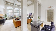 Located at 2424 E TC Jester Blvd in Houston, TX, this single-floor space presents an impressive combination of architectural elegance and functional design. The layout integrates a living room and dining area within a generously proportioned open floor plan finished with large-format travertine tile flooring throughout.

The most striking architectural feature is the dramatic ceiling height, accentuated by detailed coffered beadwork that adds a sense of grandeur across the entire space. Stately columns define zones within the open layout without interrupting sightlines, contributing to the expansive feel of the interior.

Natural light floods the space through a series of tall arched windows and transom windows positioned along the exterior walls, offering clear views of the surrounding greenery. A set of glass-paned French doors provides a welcoming entryway, reinforcing the bright, airy atmosphere.

The living area is anchored by a comfortable seating arrangement beneath a wall-mounted display, while the adjacent dining zone features bar-height seating alongside built-in shelving. An additional workstation area with built-in cabinetry provides functional versatility. The overall ambiance is clean, modern, and refined, with a neutral white and warm beige palette complemented by curated décor accents throughout.