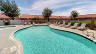 This single-story community amenity area is designed around a generously sized, freeform swimming pool with crystal-clear turquoise water, offering a resort-like atmosphere well-suited for relaxation and recreation. The pool's organic, curved shape adds a leisurely aesthetic, and an adjacent spa provides an additional option for unwinding. The expansive surrounding deck is finished in a light-toned concrete surface, providing ample space for sunbathing and socializing. Multiple lounge chairs are arranged along the pool's perimeter, along with a shaded seating area featuring an umbrella-covered table, ideal for outdoor dining or casual gatherings. The architecture framing the space is characterized by warm stucco exteriors and distinctive red clay tile rooflines, reflecting a Southwestern or Mediterranean design influence. Mature trees and desert-adapted landscaping soften the hardscape, adding natural texture and shade. A secure perimeter fence encloses the area, enhancing privacy and safety. Sliding glass doors and a dedicated entry door on the adjacent building suggest direct access to interior amenities. The overall ambiance is bright, open, and inviting, consistent with a sunny, arid climate setting.