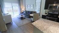 This single-floor residence showcases a thoughtfully designed open-concept layout that seamlessly connects the kitchen, dining, and living areas into one cohesive living space. The kitchen features a striking granite-topped island with distinctive veining in soft gray and white tones, complemented by dark cabinetry and a decorative tile backsplash. The island serves as a natural transition point into the dining area, where a round white table and upholstered chairs provide an intimate setting for meals.

The living area is anchored by warm hardwood-style flooring and a neutral area rug, creating a polished foundation beneath a comfortable gray sofa accented with decorative pillows. A modern glass-and-chrome coffee table adds a refined touch. Framed artwork displayed above the sofa contributes a gallery-like aesthetic to the space. A large mirror nearby enhances the sense of openness and reflects natural light throughout. Sliding glass doors flood the interior with abundant natural light while providing direct access to a private outdoor balcony, offering views of the surrounding greenery. The overall ambiance is contemporary, clean, and inviting.