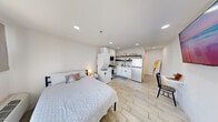 This efficiently designed 1-bedroom, 1-bathroom unit makes excellent use of its compact footprint across a single floor. The open-plan main area seamlessly integrates the sleeping, dining, and kitchen zones, all unified by light wood-look tile flooring and bright white walls that maximize the sense of space. Recessed ceiling lights illuminate the entire area evenly.

The kitchen is a standout feature, outfitted with white shaker cabinets accented by gold hardware, a marble-style tile backsplash, stainless steel gas range with over-the-range microwave, a full-size refrigerator, and an in-unit stacked washer and dryer. Open shelving above the countertops provides additional storage and display space.

The bedroom area is a defined, private room featuring a window with curtains that allow natural light to filter in, a wall-mounted HVAC unit for climate control, and a bold black entry door for security and style. A wall-mounted TV adds a modern touch to the living space.

The bathroom features a clean, minimalist design with a pedestal sink, a gold-framed vanity mirror, and a stylish multi-bulb light fixture, maintaining the modern aesthetic found throughout the unit. Every element of this space has been thoughtfully arranged to deliver functional, comfortable living in a well-appointed, contemporary setting.