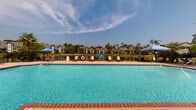This outdoor pool facility in Slidell, Louisiana offers a well-appointed communal swimming and recreation space designed for relaxed outdoor living. The centerpiece is a generously sized swimming pool with vivid turquoise water, framed by a broad concrete sun deck that provides ample space for lounging and socializing. Brick-edged coping lines the pool's perimeter, complementing the classic red brick construction of the adjacent poolside structure, which features multi-pane windows and a covered overhang supported by substantial brick columns with black metal railings.

The surrounding deck is furnished with reclining lounge chairs and shaded by large blue canopy umbrellas, offering comfortable spots to relax out of direct sunlight. A life ring station is mounted poolside, and posted signage provides pool rules for users. The entire pool area is enclosed by a black iron fence, ensuring security while maintaining open sightlines to the lush, mature tree line and the surrounding residential buildings beyond. The landscaping includes palm trees and manicured greenery that enhance the resort-like atmosphere. Under clear Louisiana skies, this pool area presents an inviting, practical, and well-maintained outdoor recreational amenity.