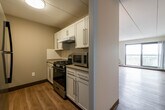 This single-floor residence offers a thoughtfully designed layout with a clean, modern aesthetic throughout. Warm hardwood-style flooring runs continuously across all areas, creating a cohesive and welcoming atmosphere.

The kitchen is fitted with white shaker-style cabinetry, light countertops, a tile backsplash, and stainless steel appliances including a gas range, microwave, and refrigerator. It connects seamlessly to the open living area, which benefits from abundant natural light streaming in through large windows and a sliding glass door.

The living area leads directly to a private balcony enclosed by a decorative railing, offering expansive elevated views of a lush, tree-lined landscape. Baseboard heating runs along the perimeter of the main living spaces, and a wall-mounted air conditioning unit provides climate control.

The bedroom is a well-sized, quiet retreat featuring its own large window with the same impressive outdoor views, a closet with bi-fold doors, and continued hardwood-style flooring. A hallway connects the rooms and provides access to additional doors and storage areas. Wall-mounted light sconces in the living space add a subtle, functional design touch. The overall ambiance is bright, clean, and move-in ready.