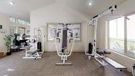 Located at 4300 Neil Rd in Reno, Nevada, this single-floor fitness center is designed to support a well-rounded workout experience. The space is divided into distinct functional zones, accommodating both cardiovascular training and strength conditioning.

The cardio area is bright and inviting, featuring soaring vaulted ceilings and an expansive wall of large windows and glass doors that flood the room with natural light and offer views of the surrounding outdoor greenery. Equipment in this zone includes elliptical trainers, a stationary recumbent bike, stair climbers, and a treadmill, all arranged to maximize space and usability.

Adjacent to the cardio area, the strength training zone houses a variety of cable and weight machines, including multi-function cable stations and leg extension/curl machines. A large wall-mounted mirror enhances the sense of space while allowing users to monitor their form. The vaulted ceiling continues into this section, maintaining the open, airy atmosphere throughout.

The facility also includes an additional section with supplementary strength equipment, a structural support column, and access to what appear to be restroom or utility facilities. Throughout the space, neutral-toned carpet flooring, recessed lighting, and clean white walls contribute to a practical yet comfortable environment well-suited for regular fitness use.