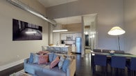 This single-floor space showcases a thoughtfully designed open-concept layout that seamlessly integrates the living, dining, and kitchen areas. The impressively tall ceilings create a dramatic sense of volume, enhanced by exposed industrial ductwork that adds an authentic loft aesthetic to the space.

The living area is anchored by a comfortable sofa adorned with colorful accent pillows, positioned beneath a striking piece of wall art. Dark hardwood flooring runs throughout, providing a rich contrast to the lighter walls and cabinetry.

The kitchen is well-appointed with white cabinetry, stainless steel appliances including a full-size refrigerator and built-in microwave, and a central island with bar seating accommodating multiple guests. Pendant lighting over the island provides focused task illumination.

Adjacent to the kitchen, a dedicated dining area features a table with upholstered chairs, ideal for more formal meals. A hallway visible toward the rear of the space leads to additional rooms, suggesting a practical and functional floor plan. The overall ambiance balances industrial character with contemporary, comfortable living.