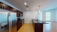 This contemporary urban unit offers a thoughtfully designed single-floor layout that blends industrial and modern aesthetics. The open-concept living and dining area is anchored by warm hardwood flooring and striking exposed concrete ceilings, which lend an industrial character to the space. Floor-to-ceiling sliding glass doors flood the living area with natural light and open to views of the surrounding cityscape, creating a bright and airy atmosphere.

The kitchen is well-appointed with rich dark-toned cabinetry, dark granite countertops, a classic white subway tile backsplash, and a full suite of stainless steel appliances including a refrigerator, electric range, and over-the-range microwave. A kitchen island with a built-in sink and pendant lighting above adds both functionality and style.

The bedroom features carpeted flooring, large windows with city views, and exposed concrete ceiling details consistent with the overall design language. Two bathrooms serve the unit, each finished with dark granite vanity tops, dark wood cabinetry, round vessel-style sinks, and large mirrors. Light tile flooring completes the clean, modern look.

A dedicated laundry area with a stacked washer and dryer provides added convenience, rounding out this well-equipped and stylish urban living space.