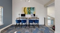 This single-floor business center is thoughtfully designed to blend functionality with contemporary style. The space is defined by soaring vaulted ceilings that create an open, airy atmosphere, complemented by oversized windows — including a striking grid-style clerestory window — that flood the interior with natural light and offer views of the surrounding neighborhood.

The main work area features two wooden desks positioned side by side against a whitewashed brick wall, each equipped with a desktop computer. Deep blue velvet accent chairs provide comfortable seating at the workstations, while a vibrant abstract painting in teal, gold, and green anchors the wall above, adding an artistic focal point. Track lighting installed along the vaulted ceiling ensures ample task illumination.

A dedicated lounge area offers a tufted white leather Barcelona-style chair alongside a glass side table and a built-in shelving nook with dark wood shelving. Decorative circular mirrors adorn the walls in both areas, reinforcing a cohesive design aesthetic. A sleek sputnik-style chandelier adds a mid-century modern touch near the entry.

The bold blue and gray patterned carpet ties the entire space together, contributing to an environment that is both professional and visually engaging.