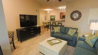 Located at 5418 Sirius Dr in Wilmington, NC, this single-floor residence offers a thoughtfully designed open-concept layout that seamlessly connects the main living areas. The living room serves as a central gathering space, featuring plush carpeting, a ceiling fan, and warm ambient lighting that creates a comfortable and inviting atmosphere. A generously sized entertainment area anchors the room, complemented by a tufted ottoman coffee table and a cozy sofa accented with decorative pillows, adding a touch of style to the functional space. Adjacent to the living room, a dining area provides an ideal spot for casual meals, furnished with a bar-height table and seating that bridges the gap between the living and kitchen spaces. The kitchen is visible from the main living area and features wood-toned cabinetry, a breakfast bar with counter seating, and dark appliances, making it both practical and accessible. Track lighting and recessed fixtures throughout ensure the space is well-lit, while the open flow between rooms enhances the sense of spaciousness. The overall design strikes a balance between comfort and practicality, making this Wilmington residence a welcoming and functional home.