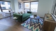This single-floor space showcases a thoughtfully designed living area that blends modern aesthetics with comfort. The room is anchored by a tufted emerald green velvet sofa adorned with an assortment of decorative pillows, creating a bold focal point. A large picture window floods the space with natural light while offering views of the surrounding urban streetscape, enhancing the open, connected feel of the interior.

Hardwood flooring runs throughout, providing a warm contrast to the cool-toned décor. A geometric patterned area rug in shades of blue, green, and grey defines the seating zone and adds visual depth. The living area is complemented by a set of circular nesting coffee tables with dark stone tops and metallic bases, alongside a sleek glass-top side table. A framed black-and-white artwork decorates the wall, contributing to the contemporary character of the space. A wall-mounted air conditioning unit ensures year-round comfort. Subtle dining furniture is visible in the background, suggesting a combined living and dining layout that maximizes the functionality of the space.