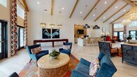 Located at 10 Shell Creek Drive in Port Royal, South Carolina, this single-floor community clubhouse delivers a bold and sophisticated coastal aesthetic throughout its thoughtfully designed interior. The entrance foyer immediately sets the tone with soaring ceilings, herringbone-patterned light wood flooring, and dramatic floor-to-ceiling windows dressed in vibrant red, navy, and white patterned drapery. An inviting reception area with a dedicated workstation provides a professional yet welcoming point of contact for visitors. The main lounge is an impressive gathering space defined by dramatic vaulted ceilings with exposed wood beam accents, statement woven globe chandeliers, and white board-and-batten wall paneling. Bold coastal artwork, a deep navy sofa, warm terracotta accent chairs, and richly layered Persian-style rugs create an inviting and visually dynamic atmosphere. An adjacent casual lounge area features plush blue velvet seating, a shuffleboard table, and a kitchenette bar with pendant lighting, ideal for social events. A dedicated conference or dining room offers an intimate setting with a substantial dark wood table, patterned wallcovering, navy drapery, and an elegant gold chandelier. Natural light flows generously throughout, with large windows offering views of the surrounding Port Royal landscape.