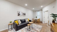 This well-appointed single-floor unit offers a clean, modern aesthetic throughout, characterized by crisp white walls, recessed lighting, and warm hardwood-style flooring that flows seamlessly from room to room. The open-concept living and dining area forms the heart of the space, where a dark leather sofa anchors the living zone alongside a colorful area rug and paired abstract wall art. The dining area, positioned near a natural-light-filled window dressed with sheer white curtains, features a round white table surrounded by natural wood chairs, creating an inviting setting for meals. The kitchen is fully equipped with stainless steel appliances — including a full-size refrigerator, range, and microwave — complemented by bright white shaker-style cabinetry, ample counter space, and a double sink. A wall-mounted mini-split HVAC unit ensures year-round climate comfort. The unit includes two bedrooms, each finished with the same warm flooring and neutral palette. One bedroom features a dedicated workspace with a desk and monitor, along with a sliding-door closet, while the second bedroom showcases a bold, colorful canvas artwork as a focal point. Both rooms offer a tranquil, restful atmosphere with generous closet storage.