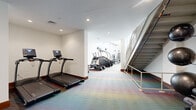 Located at 7 New York Ave NE in Washington, DC, this fitness center offers a thoughtfully designed exercise space spread across a single floor. The room is defined by clean white walls, recessed lighting, and a vibrant multicolored carpet that adds an energetic and modern aesthetic to the environment.

The space is organized to accommodate a wide variety of workout styles. The cardio section features multiple True-brand treadmills and stair climbers, providing ample options for aerobic training. A stationary cycle with an attached monitor is also present for cycling workouts. Strength training is well represented, with a cable and pulley machine, a dedicated free weights area complete with a tiered dumbbell rack and adjustable weight bench, and additional resistance equipment positioned throughout the room.

A striking open staircase with metal railings runs along one side of the space, contributing an architectural element that adds height and visual interest to the layout. In one area, the ceiling opens up to a double-height volume, enhancing the sense of openness. A water station is conveniently positioned within the space for easy access during workouts.

The layout flows naturally from one equipment zone to the next, making efficient use of the generously sized floor plan. Overall, the fitness center presents a clean, modern, and fully functional environment suited for a comprehensive exercise routine.
