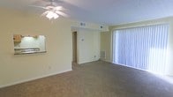This single-story unit features an open living area finished with wall-to-wall carpeting that extends throughout the main space, offering a comfortable and cohesive feel underfoot. The room is centered beneath a ceiling fan with an integrated light fixture, providing both air circulation and overhead illumination. A large window dressed with vertical blinds dominates one wall, allowing natural light to fill the space while offering privacy control. The neutral, light-toned walls contribute to a bright and airy atmosphere. A wall-mounted HVAC unit is positioned beneath the window, ensuring climate control convenience. Overhead, a ceiling vent further supports the unit's ventilation system. To one side, a partial view of a countertop suggests an adjacent kitchen area, while a doorway at the rear of the room leads to additional spaces within the unit. The overall layout is straightforward and functional, with a clean, open design that offers flexibility in furniture arrangement and use of the living space.