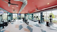 This fitness center in Gonzales, Louisiana offers a generously sized, single-floor workout facility designed to support a full range of exercise routines. The space makes an immediate visual impression with its bold red ceiling, exposed industrial-style HVAC ductwork, and teal-painted walls — a color palette that energizes the environment. Ceiling fans are distributed throughout, ensuring comfortable airflow during workouts.

The main workout floor is defined by a striking geometric patterned tile floor in shades of teal, coral, and gray, adding a modern, dynamic aesthetic underfoot. Large floor-to-ceiling windows line the perimeter, flooding the space with abundant natural light and offering views of the surrounding outdoor area, including what appears to be a pool and landscaping.

Equipment is thoughtfully arranged throughout the space. Cardio machines — including treadmills, ellipticals, and stationary bikes — are positioned along the windowed walls to take advantage of the outdoor views. A multi-function cable machine and weight bench anchor the strength training area, while a well-stocked dumbbell rack provides a full range of free weights. Wall-mounted flat-screen TVs are positioned for visibility across multiple workout zones.

A separate enclosed room, visible from the main floor, appears to serve as a dedicated stretching or group fitness area with a wood-style floor. Full-length mirrors along one wall further enhance the sense of openness and allow users to monitor their form during exercise.