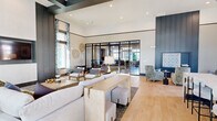 Located at 1735 Waterway Cir in Middleburg, Florida, this thoughtfully designed single-floor clubhouse offers an upscale communal experience through two well-appointed living areas connected by an open, flowing layout. The space is defined by soaring ceilings, large-format polished tile flooring in the reception and lounge zones, and wide-plank light wood flooring throughout the secondary living area, creating a warm contrast of textures.

The primary lounge features built-in banquette seating in rich slate blue, complemented by gold-toned accent tables and a sculptural multi-arm pendant light fixture. Recessed alcoves with textured wallcovering and curated artwork add depth and intimacy to the open plan. A second lounge area showcases dramatic double-height ceilings, floor-to-ceiling windows with blue botanical drapery, a large wall-mounted television framed by sleek dark paneling, and an abundance of natural light. This area also includes space for recreational games, adding a social and relaxed dimension.

A kitchenette with a white quartz countertop, vertical subway tile backsplash, dark cabinetry, and a built-in television anchors one end of the space. Glass-paneled doors open to exterior views, further enhancing the light-filled, airy ambiance that defines this modern and inviting community gathering space.