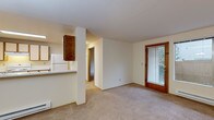 Located at 10740 NE 8th Avenue in Seattle, WA, this single-floor unit offers a practical and well-organized layout suited for comfortable everyday living. The living and dining area is finished with neutral wall-to-wall carpeting and features a chandelier overhead, a baseboard heating system, and a sliding glass door with blinds that opens to views of the surrounding greenery and outdoor fencing, allowing natural light to fill the space. A pass-through counter connects the living area to the kitchen, which is equipped with wood-framed cabinetry featuring cream-colored panels, a white electric range with an overhead ventilation hood, light-toned countertops, and a full-size refrigerator. The layout flows naturally into a hallway leading to the bedroom, which is generously proportioned with soft carpeting, a large sliding window overlooking lush landscaping, and a baseboard heater. The bathroom is finished with light tile flooring and includes a full-size soaking tub and shower combination, a toilet, and a vanity sink, with grab bars installed throughout for added accessibility and safety. Neutral tones and clean lines throughout the unit create a calm, functional atmosphere well-suited to Seattle's residential north end.