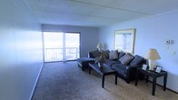 This single-floor space offers a welcoming and functional layout centered around a spacious living area. The living room is finished with wall-to-wall carpeting that adds warmth underfoot and is bathed in natural light streaming through large sliding glass doors that lead directly to a private balcony. The balcony access provides an easy indoor-outdoor connection, enhancing the sense of openness throughout the space. Vertical blinds frame the sliding doors, offering flexible light control and privacy. The living area comfortably accommodates a sectional sofa arrangement centered around a dark-finished coffee table, making it well-suited for both relaxing and entertaining. A textured ceiling adds architectural character to the room, while neutral blue-grey walls create a calm, understated backdrop. Wall space is thoughtfully utilized, with room for framed artwork that complements the overall aesthetic. A floor lamp provides supplemental ambient lighting, adding to the cozy atmosphere during evening hours. The balcony beyond the glass doors appears to overlook an open exterior area, extending the living experience outdoors. Overall, this space strikes a balance between practicality and comfort, with a layout that maximizes natural light and livability.