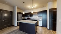 This single-floor unit showcases a thoughtfully designed open-concept layout with a cohesive contemporary aesthetic throughout. The kitchen serves as the centerpiece, featuring rich dark cabinetry that extends to the ceiling and provides generous storage space, including a distinctive lattice wine rack. Speckled granite countertops run along the perimeter and crown the sizable center island, which offers additional prep and seating space. A full suite of stainless steel appliances — including a refrigerator, electric range, built-in microwave, and dishwasher — complements the modern design. A multi-light pendant and track lighting fixture illuminate the kitchen area effectively.

Wood-look flooring extends through the kitchen and main living areas, transitioning to plush carpeting in the bedrooms. The bedroom features large windows fitted with horizontal blinds, allowing ample natural light and outdoor views. A second carpeted room with an adjacent full bathroom offers flexible use as an additional bedroom or den. The bathroom includes a tub-shower combination, wood-look flooring, granite-topped vanity, and dark-finished door hardware consistent with the unit's overall design language.