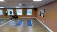 This single-floor fitness and yoga studio is thoughtfully designed to support a focused and calming workout environment. The open exercise area features smooth hardwood-style flooring, ideal for yoga, stretching, or general fitness activities, with yoga mats already in place for use. Warm terracotta-toned walls create an energizing yet grounded ambiance throughout the space. The drop ceiling is fitted with recessed panel lighting that provides even, comfortable illumination across the room. Multiple windows along one wall invite generous natural light and offer views of an outdoor pool area, connecting the interior to the surrounding outdoor environment. A wall-mounted flat-screen television is positioned for easy viewing during workouts. Along the perimeter, a rack of weighted medicine balls and additional fitness equipment is available, adding functional versatility to the studio. A towel rack with folded towels is conveniently mounted near the windows for post-workout use. A large framed wall piece adds a decorative touch, contributing to the polished and professional feel of the space. Overall, this studio offers a clean, well-equipped, and inviting setting well-suited for yoga sessions, personal training, or group fitness activities.