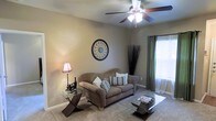 This single-floor space features a living room with a welcoming and comfortable ambiance. The room is finished with wall-to-wall carpeting and painted in warm, neutral tones that create a relaxed atmosphere. A ceiling fan with an integrated light fixture serves as a functional centerpiece overhead, providing both airflow and illumination. Natural light filters into the space through a window dressed with layered green curtains and sheer panels, adding a touch of color contrast against the soft beige walls. The living area accommodates a plush loveseat adorned with decorative striped and solid throw pillows, complemented by a glass-top coffee table with a distinctive geometric base. A matching glass side table holds a traditional table lamp, adding warm accent lighting. Decorative touches include a circular ornamental wall piece and a tall arrangement of dried grass stems, lending the room a curated, stylish feel. The overall layout is efficiently arranged, making the space feel inviting and well-organized despite its modest footprint.