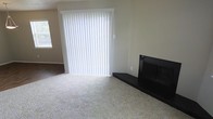 This single-story space presents a straightforward and functional living area finished with neutral-toned carpet that extends throughout the room, providing a soft, comfortable underfoot feel. The walls are painted in a warm beige tone, contributing to a calm and understated ambiance that suits a variety of décor styles.

A prominent feature of the living area is the dark-finished fireplace set against the wall, complete with a raised hearth platform finished in matching dark material. This element serves as a natural focal point and adds a touch of warmth and character to the space.

Natural light enters the room through a large sliding glass door fitted with full-length vertical blinds, which allow for adjustable light control and privacy. An additional window is visible to the side, further enhancing the brightness of the interior. The sliding door likely provides access to an outdoor area, offering a functional connection between the interior and exterior of the home.

The overall layout is open and uncluttered, making the space versatile and adaptable for various furniture arrangements. The combination of soft carpeting, neutral walls, and the fireplace creates a livable, welcoming environment throughout this single-level home.