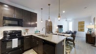 This thoughtfully designed single-floor residence showcases a seamless open-concept layout that connects the kitchen, dining, and living areas into one cohesive, flowing space. The kitchen is a standout feature, boasting rich dark cabinetry paired with light granite countertops and a classic white subway tile backsplash. A double-basin sink sits at the center of the kitchen peninsula, which is illuminated by a trio of industrial-style Edison bulb pendant lights, adding a warm, modern character to the space. Black stainless appliances, including a dishwasher and range, complement the bold cabinetry. The peninsula doubles as a breakfast bar with seating, creating a natural transition into the dining and living areas. The living room benefits from an abundance of natural light streaming through a large window and is kept comfortable by a ceiling fan overhead. Light-toned flooring runs throughout, enhancing the bright and airy ambiance of the space. The overall design strikes a balance between contemporary style and everyday functionality, making it an inviting and practical living environment.