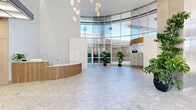 Located at 460 North Broad Street in Philadelphia, PA, this ground-floor lobby is a striking example of contemporary architectural design. The entrance area commands immediate attention with its dramatic, soaring curved glass curtain wall that stretches from floor to ceiling, flooding the interior with abundant natural light and offering open views of the surrounding cityscape. A cluster of globe pendant light fixtures descends gracefully from the high ceiling, adding a sculptural focal point above the spacious main floor.

At the heart of the entrance stands a elegantly curved reception desk clad in warm, vertically-slatted wood paneling, complementing the building's natural material palette. The flooring throughout is a polished terrazzo tile, lending a refined, upscale character to the space. Lush tropical potted plants are strategically positioned throughout the lobby and hallways, softening the architecture and adding vibrant greenery.

A bank of resident mailboxes is neatly integrated along one wall, accented by a distinctive vertical wood slat partition that provides both visual interest and subtle spatial division. The hallways extend from the main lobby, featuring warm light wood wall panels and a luminous grid ceiling that diffuses light evenly. Multiple stainless steel elevator doors line the hallway walls, providing efficient vertical access throughout the building. The overall ambiance is bright, welcoming, and polished.