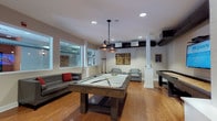 This single-floor game room is designed with entertainment and social interaction in mind, combining a modern industrial aesthetic with functional recreational amenities. The space centers around a full-size pool table with a light felt surface, positioned beneath a pair of stylish pendant lights that provide focused illumination. Adjacent to the pool table, a full-length shuffleboard table runs along one wall, accompanied by a wall-mounted flat-screen television, creating an ideal setup for group gatherings.

The flooring throughout is warm-toned hardwood, adding a classic contrast to the room's contemporary industrial design elements, which include exposed ductwork running along the ceiling. Recessed lighting supplements the pendant fixtures, ensuring the space is well-lit throughout. Structural columns with clean white casings divide the room subtly without interrupting the open layout.

Seating is thoughtfully arranged, with a leather sofa accented by red throw pillows and two modern gray upholstered armchairs with a side table, offering comfortable viewing and lounging areas. Large interior-facing windows along one wall allow visibility into adjacent spaces, contributing to an open and connected atmosphere. Decorative wall art with industrial and steampunk-inspired motifs reinforces the cohesive design theme throughout the room.