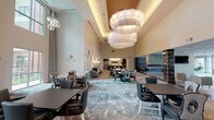 Located at 850 Imperial Blvd in Sugar Land, TX, this single-story community facility presents a sophisticated and welcoming environment designed for social gathering and daily living. The space is anchored by a grand great room with soaring ceilings adorned with striking large-scale cloud-like pendant light fixtures, creating a dramatic yet comfortable atmosphere. Patterned carpeting flows throughout the main areas, complementing the refined, contemporary décor.

The expansive dining area accommodates numerous seating configurations, from intimate round tables to larger rectangular arrangements, all set beneath generous ceiling heights that amplify the sense of openness. Large floor-to-ceiling windows and glass doors bathe the space in abundant natural light while offering views of lush outdoor greenery and a resort-style pool area, which is directly accessible from the interior.

A fully equipped commercial kitchen supports the dining area, featuring stainless steel appliances, professional-grade range with hood, built-in ovens, and a large center island with counter seating. Adjacent lounge areas provide comfortable seating clusters with upholstered chairs and sofas arranged for relaxed conversation. A bar area and decorative stone accent columns add architectural character throughout. The grand entry lobby features a tufted accent wall and direct views to the outdoor courtyard.