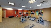This fitness center is designed to accommodate a well-rounded workout experience within a single-level layout. The space is organized into distinct functional zones, beginning with a cardio area outfitted with multiple Precor treadmills, elliptical trainers, and stationary bikes, all positioned on protective floor mats. Colorful pop-art panels adorn the warm-toned walls, adding visual energy to the workout environment.

A dedicated free weights section features a structured dumbbell rack with an accompanying mirror wall, allowing users to monitor their form during strength training. A flat workout bench and exercise mat area provide additional space for floor-based exercises and stretching routines.

The space is finished with a drop tile ceiling equipped with recessed and fluorescent panel lighting, ensuring consistent illumination throughout. Patterned carpet tile flooring runs throughout, offering both durability and comfort underfoot. Natural light filters in through multiple windows fitted with horizontal blinds along one wall. A wall-mounted flat-screen television, a water fountain, and an exercise and calorie reference chart are among the practical amenities present. Bold terracotta and golden-yellow accent walls unify the space, contributing to an energetic and motivating atmosphere.