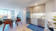 This thoughtfully designed single-floor residence offers a cohesive open-concept layout that seamlessly connects the kitchen, dining, and living areas. The contemporary kitchen is outfitted with light wood cabinetry extending to the ceiling, white subway tile backsplash, gleaming white countertops, and a full suite of stainless steel appliances including a refrigerator, dishwasher, and range. Under-cabinet lighting enhances the workspace, while light hardwood flooring flows throughout the common areas, contributing to a bright and airy atmosphere.

The dining area sits adjacent to the kitchen, providing an intimate setting for everyday meals. The living room is generously proportioned, anchored by large floor-to-ceiling windows that flood the space with natural light and offer expansive urban views of the surrounding cityscape.

The bedroom continues the modern aesthetic with its own floor-to-ceiling sliding glass door that frames striking views of the city's historic architecture. Light-toned walls and soft carpeting create a calm, restful environment.

The full bathroom features large-format tile flooring, a combined shower and bathtub with a glass slider, a light wood vanity with an undermount sink, and recessed lighting, completing this well-appointed residence.