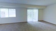 This two-floor space offers a clean, functional layout designed for comfortable everyday living. The main living area is finished with wall-to-wall carpeting in a neutral tone, creating a warm and inviting atmosphere. A prominent sliding glass door fitted with vertical blinds serves as a centerpiece of the room, flooding the interior with natural light while providing direct views of the lush greenery outside. A secondary side window further enhances the brightness of the space, ensuring the area remains airy throughout the day. The walls are finished in a soft, neutral palette, complemented by classic wood baseboards that run throughout. A textured ceiling adds a subtle architectural detail overhead. The open and uncluttered layout allows for versatile furniture arrangement, accommodating a range of living configurations. The space spans two floors, offering distinct zones for living and other functions, with the overall design emphasizing simplicity, natural light, and a connection to the outdoors. The combination of neutral finishes and abundant daylight creates a calm, welcoming ambiance suited to a variety of lifestyles.
