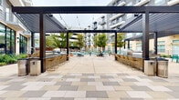 This expansive outdoor amenity space is designed for resort-style living, offering a thoughtfully curated environment for relaxation and social gathering. At its centerpiece sits a generously sized swimming pool with clear, blue-tinted water, complemented by an adjacent spa, both framed by rows of teal-colored lounge chairs along the pool deck. The surrounding pavement features a stylish geometric pattern of alternating light and neutral stone tiles that adds visual interest throughout the courtyard.

A striking modern pergola anchors the entertaining zone, constructed with sleek dark metal beams and accented by strung bistro lights overhead. On either side of the pergola, bar-height counters with gold wire stools provide ample seating for gatherings, while nearby outdoor grilling stations offer stainless steel cooking equipment ideal for social events.

Colorful patio furniture — including vibrant chairs in purple, teal, and white — is arranged around raised planting beds featuring ornamental trees and flowering plants, adding lively greenery to the space. Lush ground-level landscaping with native shrubs and grasses borders the building's exterior, which showcases a contemporary facade blending brick, metal panel, and large floor-to-ceiling windows. The entire courtyard is enclosed by the surrounding multi-story building, creating an intimate yet open-air atmosphere.