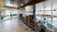 This elegant single-floor space presents a refined blend of modern and warm design elements throughout. Large-format stone-look tile flooring runs seamlessly across the entire floor, lending a sense of openness and continuity. Rich, dark wood paneling lines the ceilings, adding warmth and a distinctive architectural character to the space.

The lounge area is thoughtfully appointed with high-back leather and plaid upholstered chairs arranged over a patterned teal and neutral area rug, creating an inviting and stylish seating arrangement. A glass railing borders an open overlook, providing visual connection to an adjacent space below illuminated by pendant lighting.

A dedicated refreshment station features sleek wood cabinetry with dark countertops, a coffee and beverage setup, and decorative white orchid arrangements that add a polished finishing touch. An accent wall with bold geometric wood paneling in contrasting tones serves as a striking design feature nearby.

The elevator lobby area is distinguished by a textured botanical-patterned wallcovering in muted tones, a curated piece of abstract wall art, and stainless steel elevator doors. Recessed and surface-mounted lighting throughout ensures the space is well-lit while complementing its upscale ambiance. An adjacent glass-walled room visible from the lounge suggests additional meeting or workspace functionality.