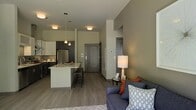 This single-floor space showcases a thoughtfully designed open-concept layout that connects the kitchen, dining, and living areas into one cohesive environment. The kitchen is appointed with dark cabinetry, a stainless steel refrigerator, a tile backsplash, and a central island with a light-colored countertop that provides both workspace and casual seating via bar-height stools. Pendant lighting above the island adds a modern, decorative touch alongside recessed ceiling fixtures that illuminate the space consistently throughout.

The living area flows naturally from the kitchen, featuring neutral olive-green walls that lend a calm, contemporary ambiance. Warm-toned laminate hardwood flooring runs throughout, unifying the open layout. A small accent table with a lamp adds warmth to the living space beside comfortable seating.

A structural column subtly defines the transition between the kitchen and living zones without disrupting the open feel. The entryway, visible from the main living area, features a modern paneled door in a muted gray tone. Elevated ceiling heights contribute to the overall sense of openness and volume within the space.