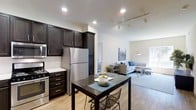This single-floor, two-bedroom, two-bathroom residence is designed with an open-concept layout that seamlessly connects the kitchen, dining, and living areas. The kitchen is outfitted with rich, dark-stained cabinetry that extends to the ceiling for ample storage, stainless steel appliances including a full-size refrigerator and range, and a classic white subway tile backsplash that adds a clean contrast to the darker tones. Light-toned hardwood-style flooring runs throughout the main living areas, contributing warmth and visual continuity across the space. A dining area sits adjacent to the kitchen, offering a natural transition point between the cooking and living zones. The living room benefits from recessed and track lighting overhead, creating a bright and well-lit environment. Neutral wall tones and generous ceiling height lend an open, airy feel to the entire floor plan. The overall ambiance is modern and functional, balancing practical storage and living needs with a cohesive, contemporary aesthetic suited for everyday comfort.