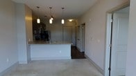 This single-floor residence showcases a clean, modern design with an open-concept layout that connects the kitchen and living areas seamlessly. The kitchen serves as a focal point, featuring a spacious island with a granite or stone-composite countertop and a sleek undermount sink. Crisp white cabinetry lines the kitchen, complemented by stainless steel appliances, including a microwave, that lend a polished, contemporary feel. Three pendant lights hang in a row above the island, providing both functional task lighting and a stylish visual anchor. Dark hardwood flooring runs through the kitchen and hallway, creating a striking contrast with the light-toned carpet that defines the living area. A neutral color palette of soft grays and whites carries throughout, lending the space a calm, cohesive ambiance. The hallway leads to additional rooms, with multiple doors suggesting well-defined private spaces tucked away from the open living zone. The layout is practical and efficiently designed, making excellent use of the single floor plan. Overall, the space balances modern aesthetics with functional living, making it a well-appointed and inviting residence.