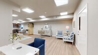 This thoughtfully designed single-floor space functions as a full-service salon and relaxation lounge, combining functional salon amenities with a welcoming, coastal-inspired interior aesthetic. Light wood-look plank flooring runs throughout, lending warmth and visual continuity across all areas.

The main lounge area features a generously sized navy blue sofa accented by rattan side tables and table lamps, complemented by abstract coastal artwork in gold and blue tones hung above. A coordinating blue and white patterned area rug anchors the seating arrangement, adding texture and color.

The salon station area is equipped with a professional hydraulic styling chair positioned before a round decorative mirror atop a white lattice-patterned credenza. A striking gold branch-style room divider elegantly separates zones within the open layout. White-framed accent chairs with blue patterned upholstery are arranged throughout for guest comfort.

The reception area includes a wood-toned desk with a blue accent chair and coastal artwork, welcoming visitors near the entry door. A dedicated utility area features dark wood cabinetry, a countertop workspace, sink, and a hair dryer station with natural light from a window.

Flush skylight panels and recessed lighting illuminate the entire space evenly, creating a bright, clean, and professional ambiance throughout.