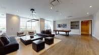 Located at 65 Bay St in Jersey City, NJ, this single-floor amenity space is thoughtfully designed to provide residents with a versatile and engaging recreational environment. The interiors are finished with wide-plank hardwood flooring, neutral-toned walls, and recessed lighting throughout, creating a clean, contemporary aesthetic.

The expansive main lounge serves as the centerpiece of the space, featuring a generous seating arrangement with a large sectional sofa, leather ottomans, and a rectangular pendant light fixture overhead. Built-in shelving, a wall-mounted flat-screen TV, and abundant natural light from oversized windows contribute to an inviting and functional atmosphere.

The game area is well-equipped, offering a classic billiards table and a full-size ping-pong table, both positioned within open, unobstructed zones that allow for comfortable play. A foosball table is also visible within the larger open area, adding to the variety of entertainment options available.

A separate children's playroom is bright and welcoming, carpeted for comfort, and outfitted with toy storage, child-sized furniture, a wall-mounted TV, and a statement gold pendant light. Windows allow ample natural light into this dedicated space, making it both practical and cheerful for younger residents.