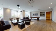 Located at 65 Bay St in Jersey City, NJ, this single-floor amenity space is thoughtfully designed to provide residents with a versatile and engaging recreational environment. The interiors are finished with wide-plank hardwood flooring, neutral-toned walls, and recessed lighting throughout, creating a clean, contemporary aesthetic.

The expansive main lounge serves as the centerpiece of the space, featuring a generous seating arrangement with a large sectional sofa, leather ottomans, and a rectangular pendant light fixture overhead. Built-in shelving, a wall-mounted flat-screen TV, and abundant natural light from oversized windows contribute to an inviting and functional atmosphere.

The game area is well-equipped, offering a classic billiards table and a full-size ping-pong table, both positioned within open, unobstructed zones that allow for comfortable play. A foosball table is also visible within the larger open area, adding to the variety of entertainment options available.

A separate children's playroom is bright and welcoming, carpeted for comfort, and outfitted with toy storage, child-sized furniture, a wall-mounted TV, and a statement gold pendant light. Windows allow ample natural light into this dedicated space, making it both practical and cheerful for younger residents.