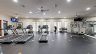 This fitness center in Annapolis, MD offers a single, generously sized exercise room designed to support a well-rounded workout experience. The space features durable speckled rubber flooring throughout, providing a safe and functional surface for all types of exercise. Large, multi-pane windows line the curved walls, flooding the room with natural light and offering views to the outdoors, while ceiling fans and recessed lighting ensure a comfortable and well-lit environment.

The cardio area is equipped with multiple treadmills and an elliptical trainer, alongside an upright stationary bike, catering to a variety of cardiovascular training preferences. A multi-tier dumbbell rack stocked with a full range of free weights provides ample options for strength training. The room also features dedicated strength machines, including a cable/pulley system and selectorized weight machines, as well as adjustable benches for versatile lifting exercises.

Full-length mirrors along one wall visually expand the space and allow for proper form checks during workouts. A wall-mounted television adds an entertainment option for users. The entrance area connects conveniently to an exterior stairway. Overall, the fitness center presents a clean, bright, and comprehensively equipped environment suitable for both cardio and strength training routines.