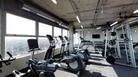 The fitness center at 800 City Tower Apartments is a well-equipped, modern facility designed to meet a range of workout needs. The space features an industrial-inspired aesthetic, with exposed concrete columns, visible ductwork, and an open ceiling that lend it a contemporary, urban character. Overhead fluorescent lighting ensures the area is well-lit throughout.

A row of cardio machines, including ellipticals and treadmills, lines the perimeter of the space, positioned directly alongside expansive floor-to-ceiling windows. These large windows allow abundant natural light to flood the room while offering sweeping panoramic views of the surrounding cityscape, providing an engaging backdrop during workouts.

On the opposite end of the fitness center, a well-stocked dumbbell rack holds a full range of free weights, catering to strength training needs. A wall-mounted flat-screen television is positioned nearby, adding entertainment options for users. The flooring is finished with durable, textured gym-appropriate material suited for high-traffic use.

Overall, the fitness center combines functional design with an energizing atmosphere, making it a practical and visually appealing amenity within 800 City Tower Apartments.