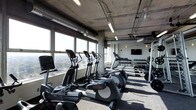 The fitness center at 800 City Tower Apartments is a well-equipped, modern facility designed to meet a range of workout needs. The space features an industrial-inspired aesthetic, with exposed concrete columns, visible ductwork, and an open ceiling that lend it a contemporary, urban character. Overhead fluorescent lighting ensures the area is well-lit throughout.

A row of cardio machines, including ellipticals and treadmills, lines the perimeter of the space, positioned directly alongside expansive floor-to-ceiling windows. These large windows allow abundant natural light to flood the room while offering sweeping panoramic views of the surrounding cityscape, providing an engaging backdrop during workouts.

On the opposite end of the fitness center, a well-stocked dumbbell rack holds a full range of free weights, catering to strength training needs. A wall-mounted flat-screen television is positioned nearby, adding entertainment options for users. The flooring is finished with durable, textured gym-appropriate material suited for high-traffic use.

Overall, the fitness center combines functional design with an energizing atmosphere, making it a practical and visually appealing amenity within 800 City Tower Apartments.
