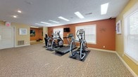 The fitness center at 1900 S Oakdale Dr in Bloomington, Indiana, is a single-floor facility designed to accommodate a range of workout needs. The space is divided into two interconnected rooms, each serving a distinct function while sharing a cohesive aesthetic defined by warm-toned walls, patterned carpeting, and textured ceilings with recessed and flush-panel lighting throughout.

The main cardio room features warm terracotta-colored walls and is equipped with multiple treadmills and elliptical machines arranged to allow comfortable spacing between users. Natural light enters through windows fitted with horizontal blinds, and a wall-mounted flat-screen television provides entertainment during workouts. The room offers ample open floor space for stretching or additional exercises.

The adjoining strength training room features softer yellow walls and houses a multi-function cable machine, a full dumbbell rack ranging across multiple weight increments, an adjustable workout bench, and additional free weights. A large mirror on one wall visually expands the space and allows users to monitor their form. Recessed lighting provides consistent illumination throughout, and colorful wall art adds a lively touch to both rooms.

Together, the two rooms form a comprehensive and functional fitness facility well-suited for varied training routines.