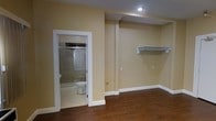 This single-floor space offers a functional and straightforward layout designed for efficient use. The main room features rich, dark hardwood-style flooring that contrasts cleanly against the warm beige walls and crisp white baseboards and trim, lending the space a polished, cohesive appearance. Recessed lighting in the ceiling provides focused illumination, while the neutral wall tones help maintain a bright and open atmosphere. A wall-mounted closet shelf with a hanging rod is built directly into one wall, offering convenient open storage without occupying additional floor space. The room's structural beam detail adds an architectural element to the ceiling, subtly defining the space. An attached bathroom is accessible through a white-trimmed doorway and includes a glass-enclosed shower-tub combination, a toilet, and tile flooring, providing all essential amenities in a compact footprint. The clean finishes throughout, including the white fixtures and consistent trim work, give the space a well-maintained and move-in-ready feel. Overall, the space is practical and neatly finished, suited for use as a bedroom or studio-style living area.