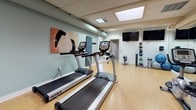 This single-level fitness center is designed to accommodate a full range of workout activities within a clean, functional layout. The space features light wood flooring throughout, which contributes to a bright and energetic atmosphere, complemented by soft sage-green walls and a drop tile ceiling with recessed and skylight-style lighting panels that keep the room well-illuminated.

Cardio equipment is prominently featured and includes multiple TRUE-brand treadmills, elliptical trainers, upright and recumbent stationary bikes, all arranged in organized rows to maximize usability. A dedicated strength training area offers a cable machine, adjustable bench, and free weight rack. Stability balls, a medicine ball tower, and additional accessories are neatly arranged along the walls.

Wall-length mirrors run along one side of the room, visually expanding the space and allowing users to monitor their form. Multiple flat-screen televisions are mounted on the walls, providing entertainment during workouts. A window at one end of the room allows natural light to enter. The overall ambiance is practical and well-maintained, making this fitness center a fully functional facility suited for a variety of exercise routines.