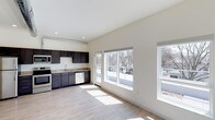 This modern studio offers a smartly designed single-floor layout that maximizes every square foot. The open-concept living and kitchen area is bathed in natural light, thanks to an expansive set of large windows and a sliding glass door that frame outdoor views of surrounding trees and a private balcony or terrace beyond. Light-toned hardwood-style flooring runs throughout, enhancing the sense of openness and warmth. The kitchen is well-appointed with rich dark cabinetry, granite-style countertops, stainless steel appliances — including a range, microwave, and dishwasher — and a single-basin sink, creating a functional and stylish cooking space. Recessed lighting accents the clean white ceilings, while an exposed conduit detail adds an industrial design touch. The neutral, light-colored walls provide a versatile backdrop that complements a variety of interior styles. The full bathroom completes this efficient yet refined layout. Overall, the space strikes a balance between contemporary aesthetics and practical living, making it an ideal setting for those seeking a low-maintenance, well-lit, and thoughtfully designed home.