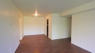 This single-floor space presents a clean, open layout characterized by dark hardwood-style flooring that runs continuously throughout the main living area, creating a warm and cohesive aesthetic. The neutral white walls provide a bright, versatile backdrop suited to a variety of uses and décor styles. A flush-mount ceiling light fixture illuminates the space, contributing to its simple, understated design. The room benefits from a generous sense of openness, with a partial wall or architectural divider adding subtle definition without fully enclosing the space. Visible through an opening in the background is an adjacent area — likely a closet or secondary room — which hints at additional storage or functional space beyond the main living area. White baseboards provide a clean contrast against the dark flooring, adding a finishing detail to the overall presentation. The layout appears straightforward and practical, offering flexibility in how the space can be arranged and utilized. Overall, the space conveys a tidy, move-in-ready quality with a neutral palette that serves as a solid foundation for personalization.
